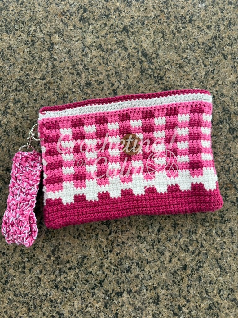 Pink fuchsia and white gingham plaid crochet zippered pouch with a sparkly white top border, a round dragonfly "choose joy" tag, and a matching marled pink wrist strap laying flat on a granite countertop.