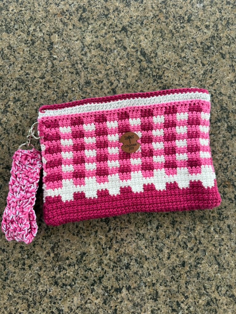 Pink fuchsia and white gingham plaid crochet zippered pouch with a sparkly white top border, a round dragonfly "choose joy" tag, and a matching marled pink wrist strap laying flat on a granite countertop.