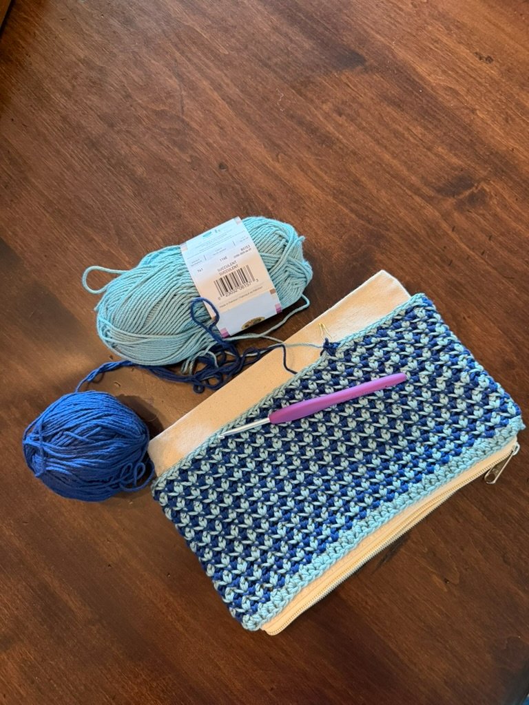 In progress shot of Navy blue and light green geometric checkered crochet zippered pouch sitting on a wood surface with a crochet hook and two skeins of yarn. The zippered canvas pouch is shown underneath the crochet design