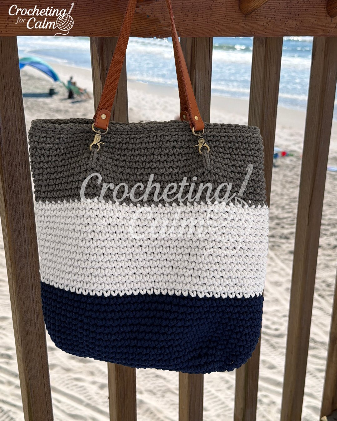 A handmade color-block crochet beach bag hanging from a wooden deck railing with a sandy beach and ocean waves in the background. The large tote bag features horizontal striped sections in navy blue, cream, and taupe gray, finished with brown leather shoulder straps attached by metal clips.