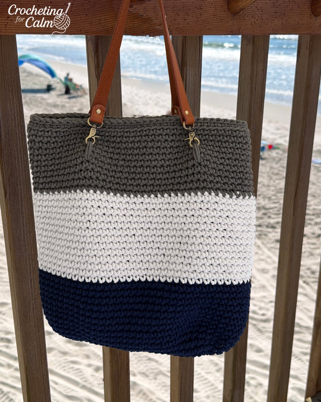 A handmade color-block crochet beach bag hanging from a wooden deck railing with a sandy beach and ocean waves in the background. The large tote bag features horizontal striped sections in navy blue, cream, and taupe gray, finished with brown leather shoulder straps attached by metal clips.
