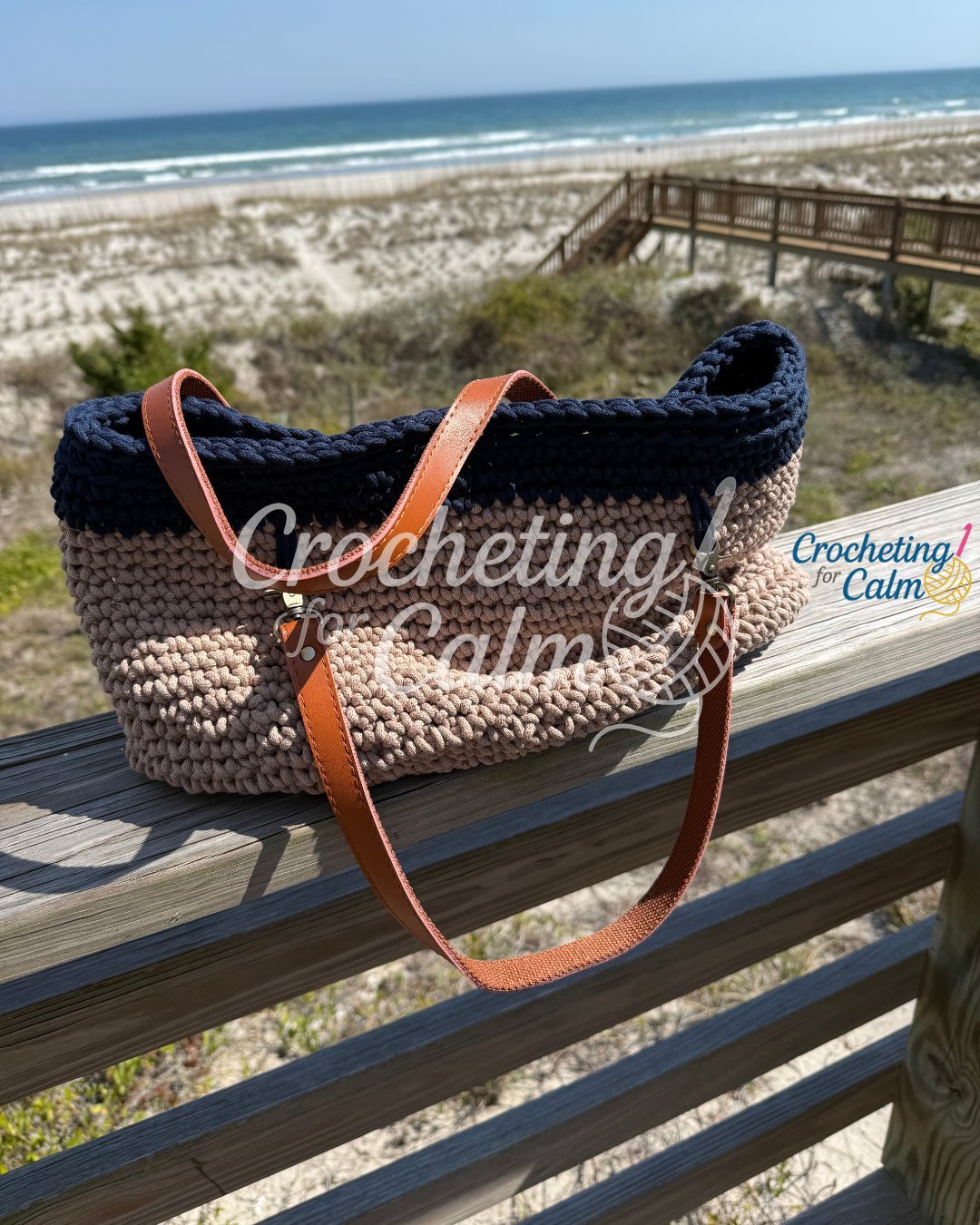 Dark navy blue and tan crochet coastal tote bag with tan leather handles, resting upright on a weathered wooden balcony railing with a sandy beach and ocean waves blurred in the background.