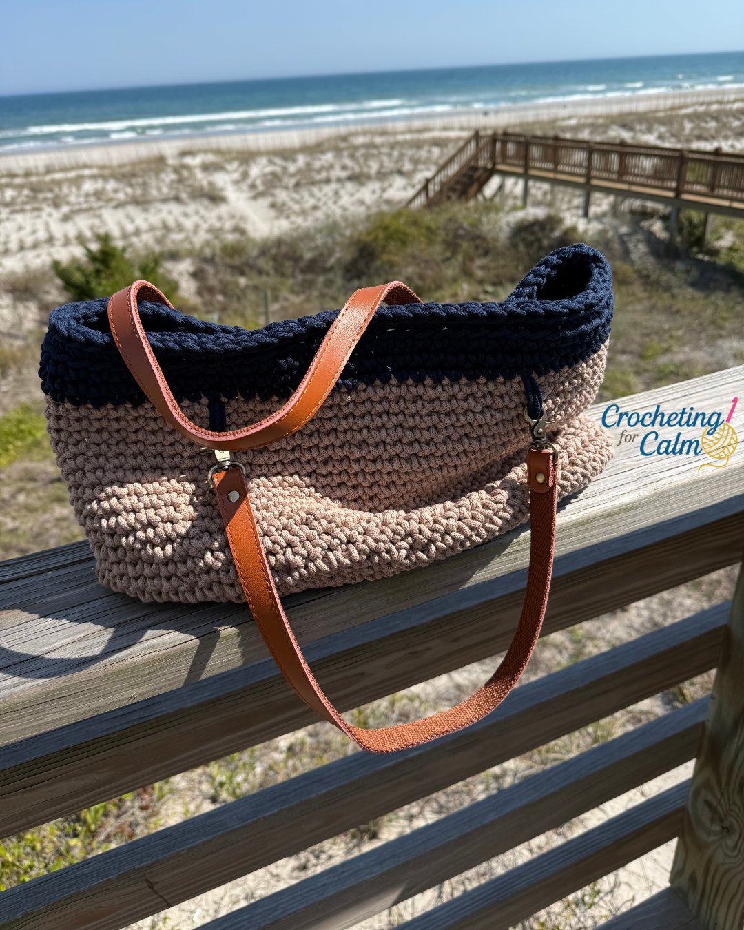 Dark navy blue and tan crochet coastal tote bag with tan leather handles, resting upright on a weathered wooden balcony railing with a sandy beach and ocean waves blurred in the background.