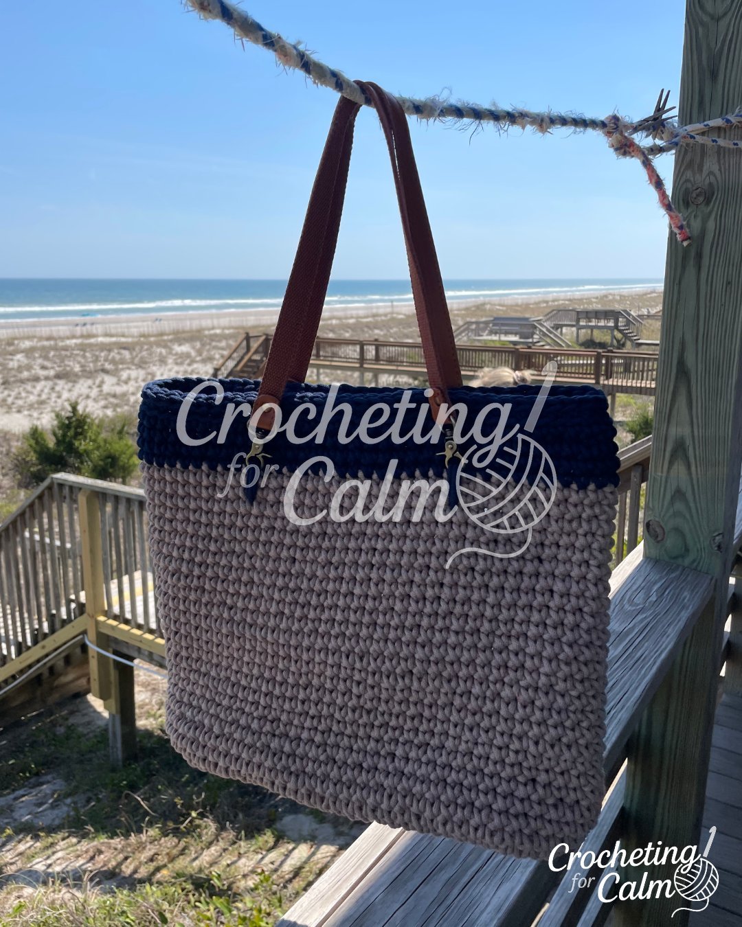 A handmade crochet beach bag hanging on a clothesline at a beach house balcony overlooking the ocean. The tote bag features a textured tan body, a dark navy blue upper border, and brown leather shoulder straps. Text overlay reads "beach bag" alongside the Crocheting for Calm logo.