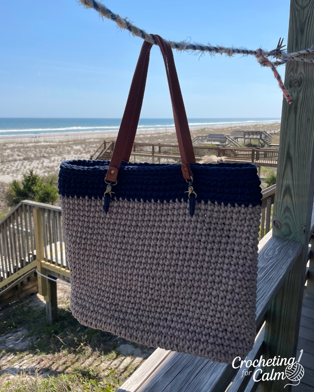 A handmade crochet beach bag hanging on a clothesline at a beach house balcony overlooking the ocean. The tote bag features a textured tan body, a dark navy blue upper border, and brown leather shoulder straps. Text overlay reads "beach bag" alongside the Crocheting for Calm logo.