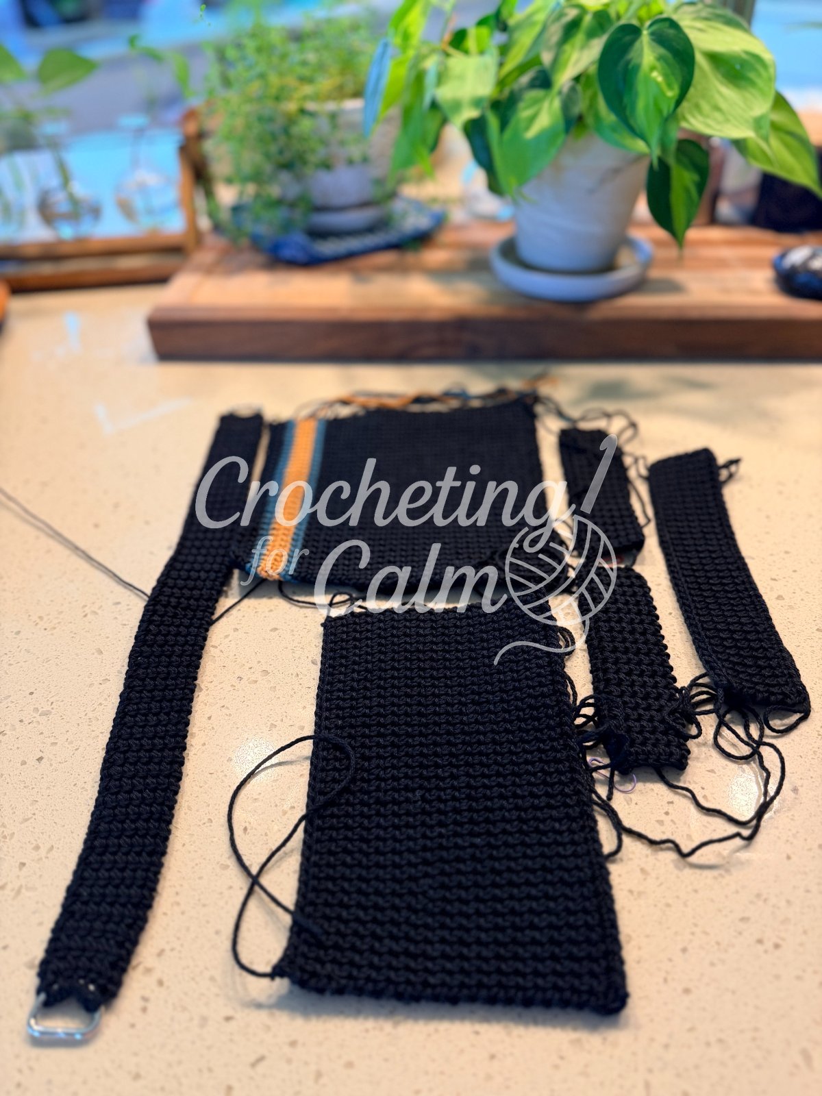 lat lay of crocheted black bag pieces prepped for assembly on a countertop with a strap, side gussets, and front/back panels featuring a teal and gold striped accent.