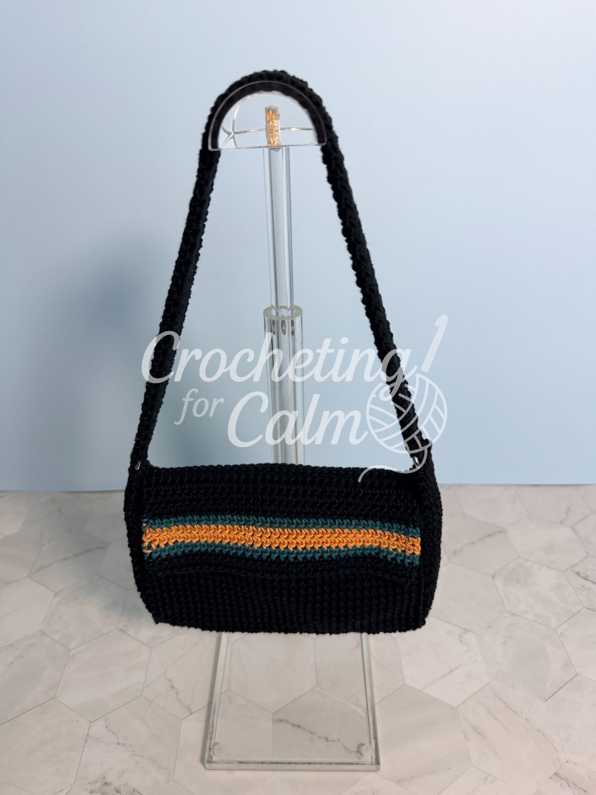 Finished black crochet shoulder bag with a teal and gold striped flap and matching wide crochet strap, hanging on a clear acrylic display stand against a light blue wall.