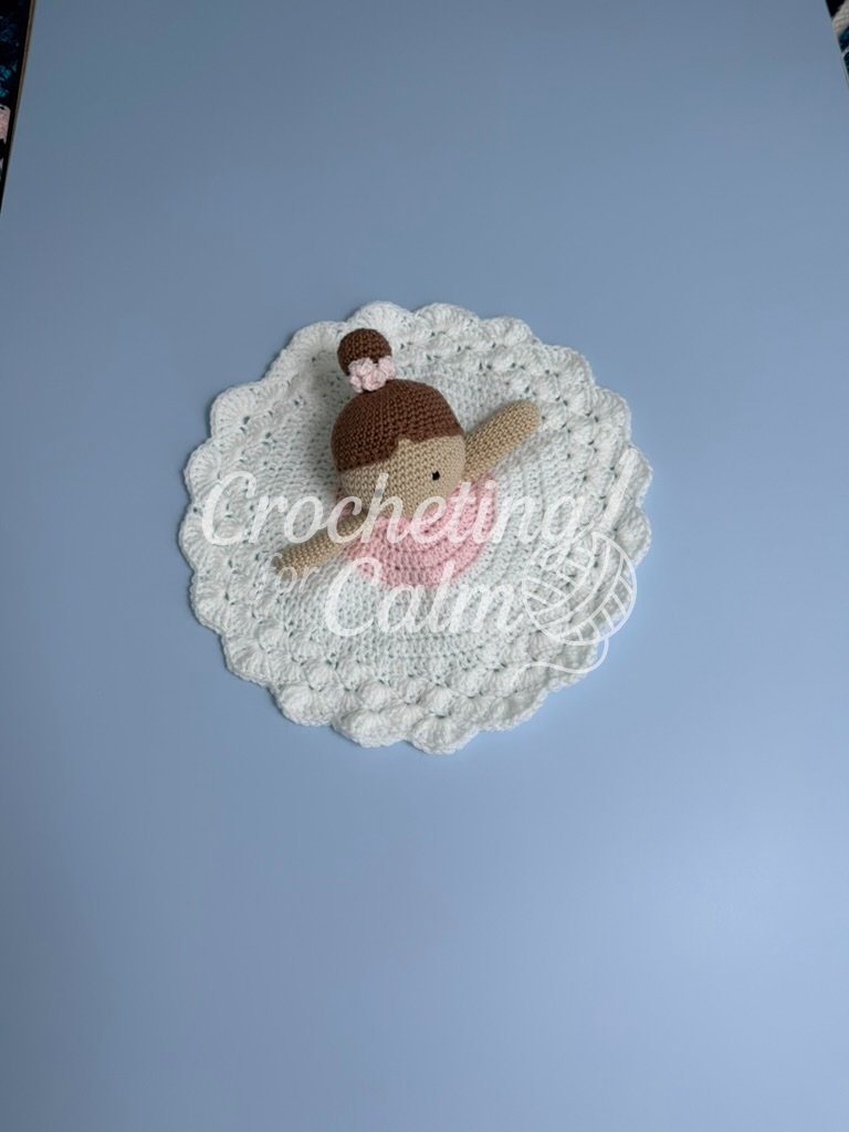 Crochet ballerina lovey featuring an amigurumi doll with brown hair in a high bun with a pink scrunchie, resting atop a circular pink and white textured blanket body with scalloped edges against a pale blue background.