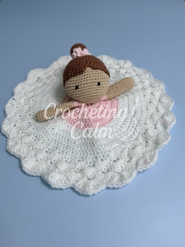 Crochet ballerina lovey featuring an amigurumi doll with brown hair in a high bun with a pink scrunchie, resting atop a circular pink and white textured blanket body with scalloped edges against a pale blue background.