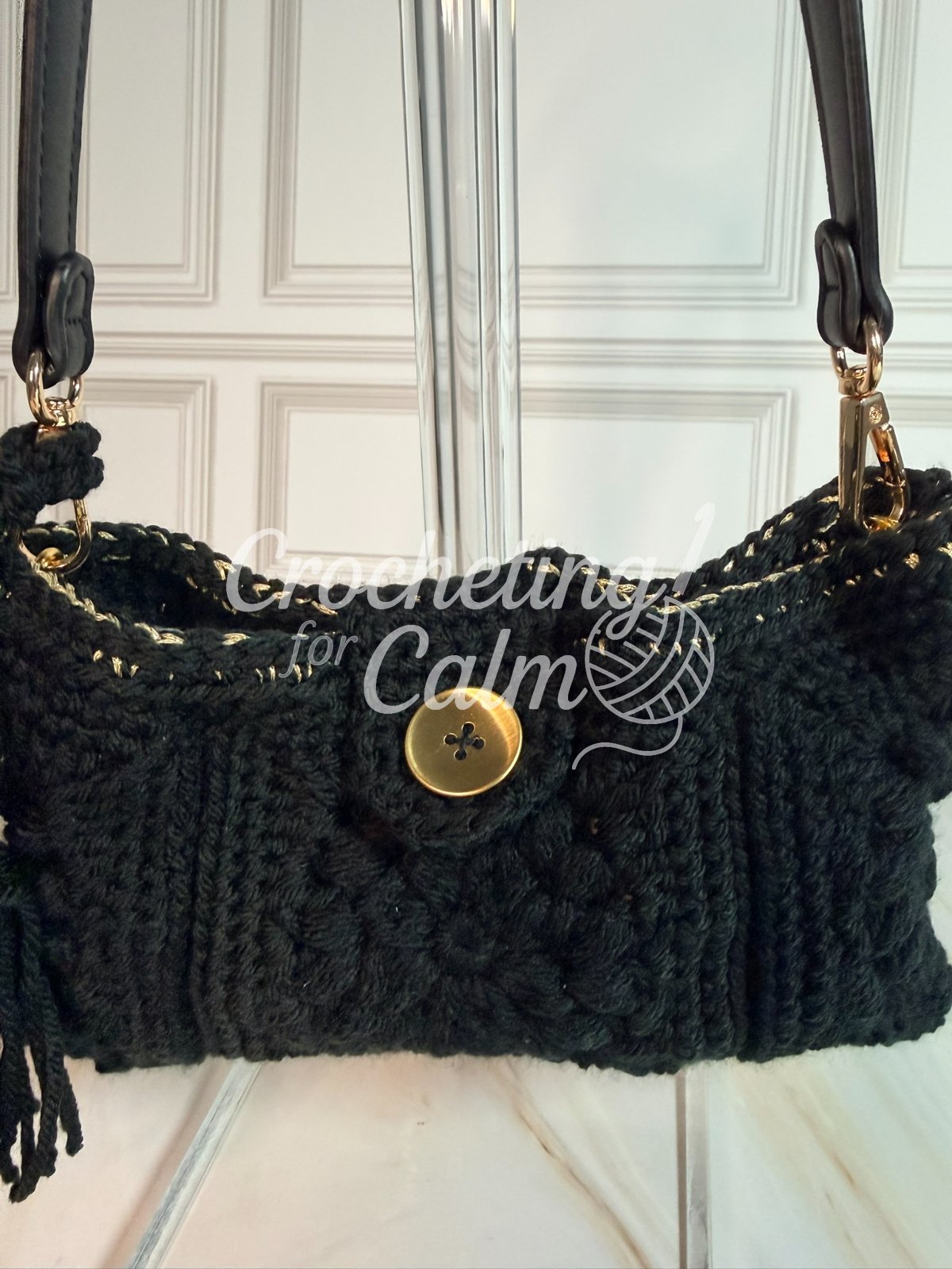 Close-up front view of a black textured crochet shoulder bag highlighting the center gold button closure, metallic gold top edge stitching, and gold strap hardware.