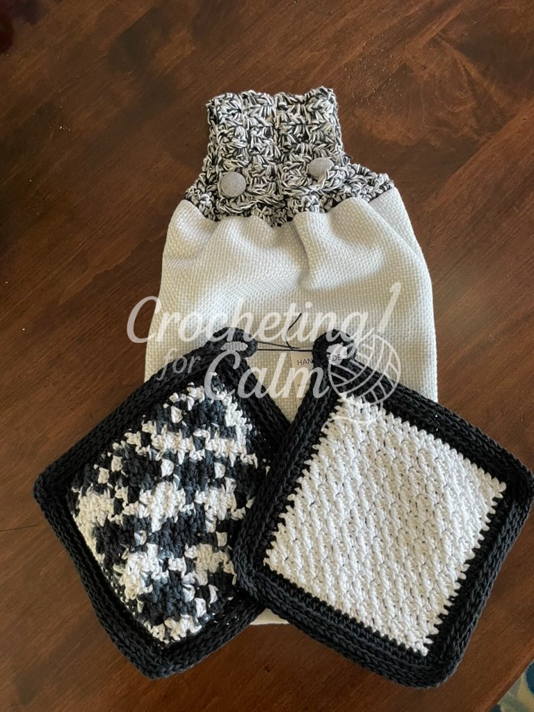 Black and white textured crochet towel topper on a white waffle-weave towel, paired with two coordinating black and white crochet potholders with hanging loops on a dark wood surface.