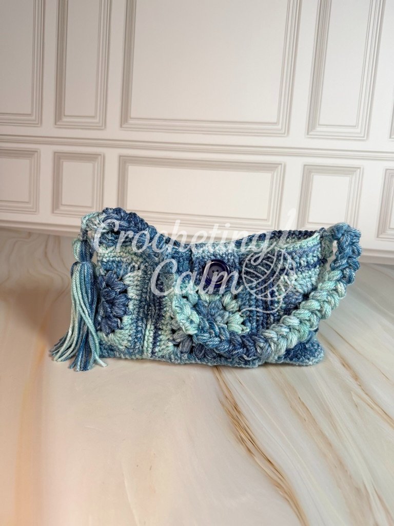Blue, light blue, and cream granny square crochet shoulder bag with a dark blue button closure and side tassel accent, resting on a marble surface.