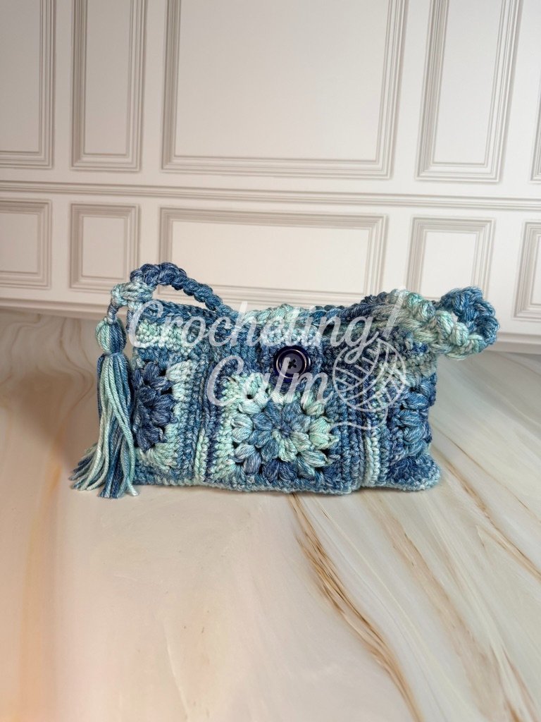 Front-facing view of a blue and cream floral granny square crochet shoulder bag with a dark blue center button and twisted strap, resting on a marble surface.