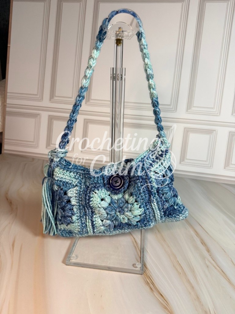 Blue, light blue, and cream granny square crochet shoulder bag with a braided shoulder strap and yarn tassel, displayed hanging on a clear acrylic stand