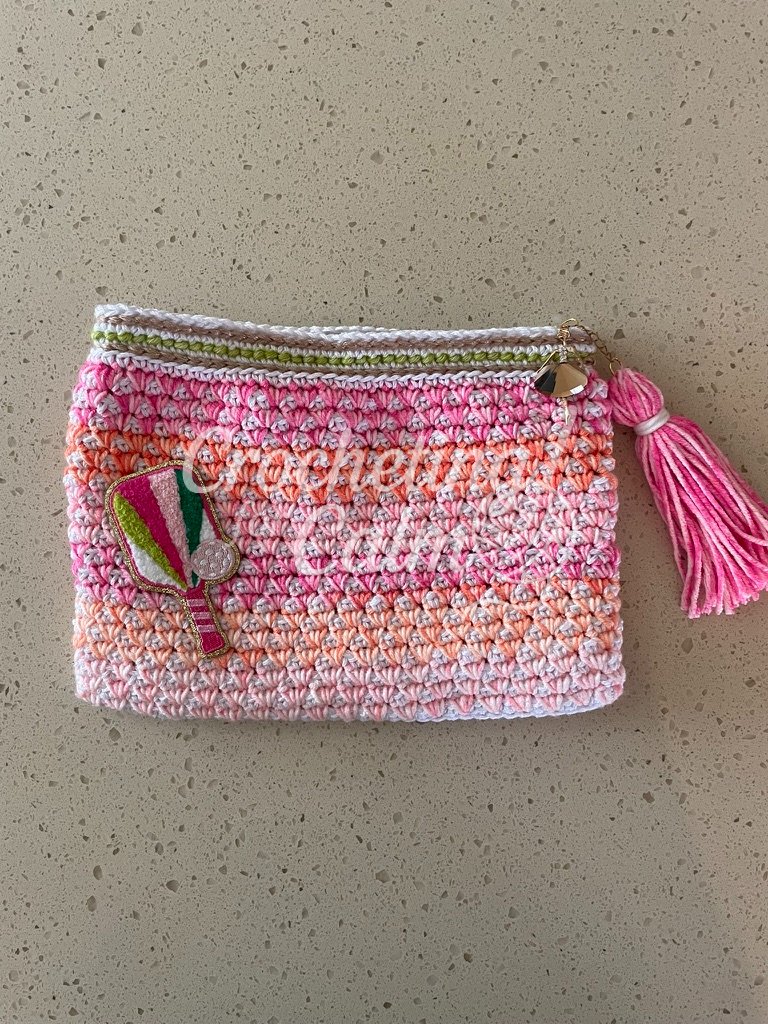 Pink, orange, and white striped crochet pouch laying flat on a neutral countertop, featuring an embroidered pickleball paddle patch on the lower left and a pink yarn tassel with a gold ballerina charm attached to the zipper.