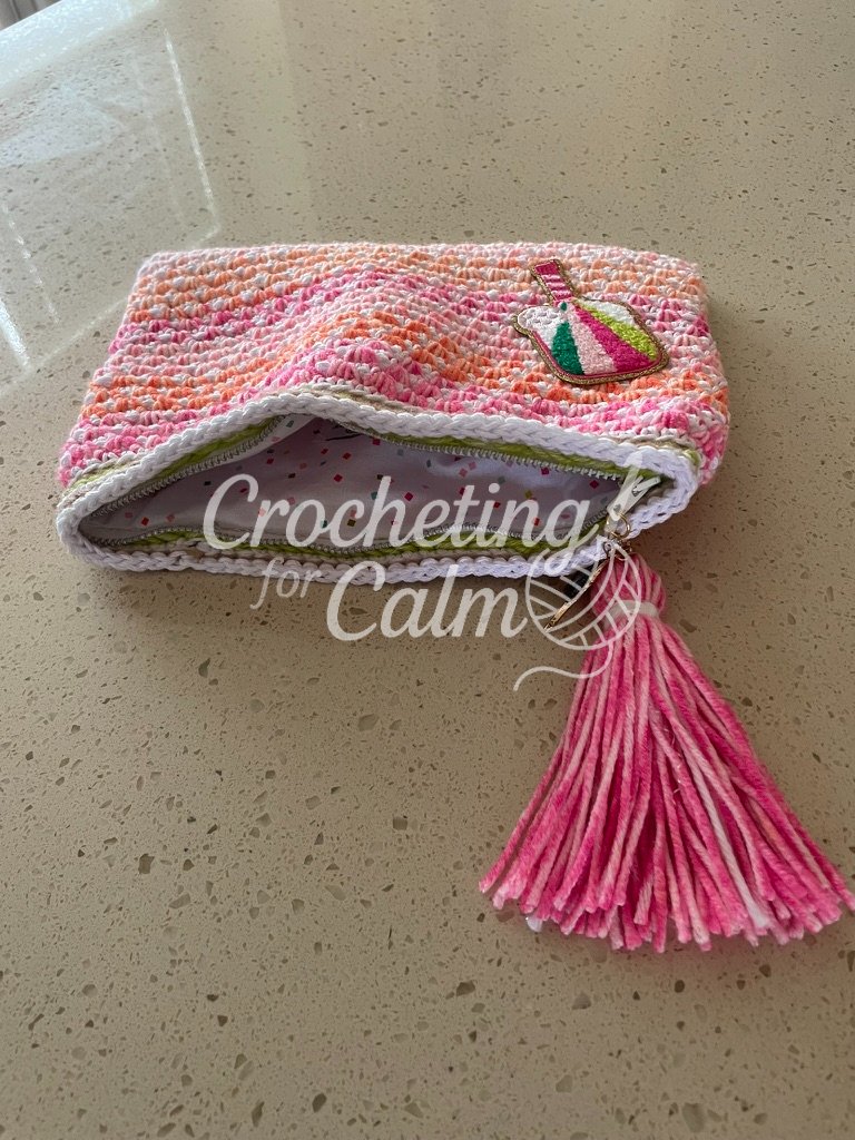 Pink, orange, and white striped crochet zippered pouch slightly unzipped to reveal a white confetti-dot fabric lining, with a pink yarn tassel and pickleball paddle patch resting on a speckled countertop.