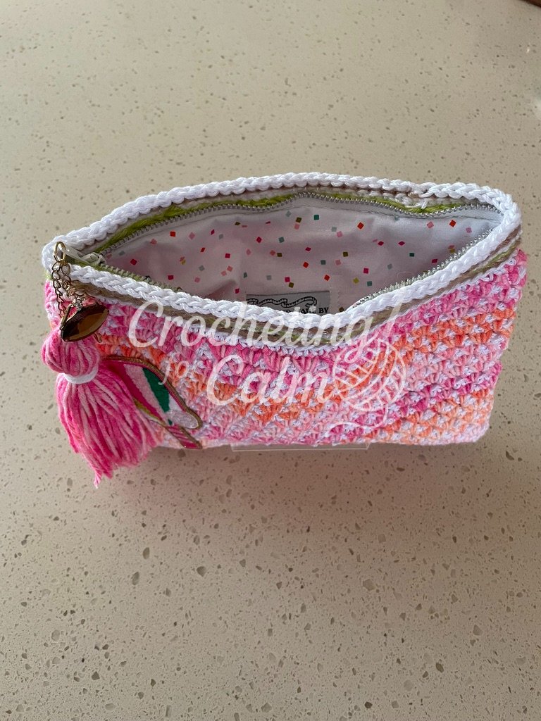Open top-down view of a pink and orange crochet pouch showing the white confetti-print fabric lining and interior maker label, with a pink yarn tassel hanging from the gold zipper.