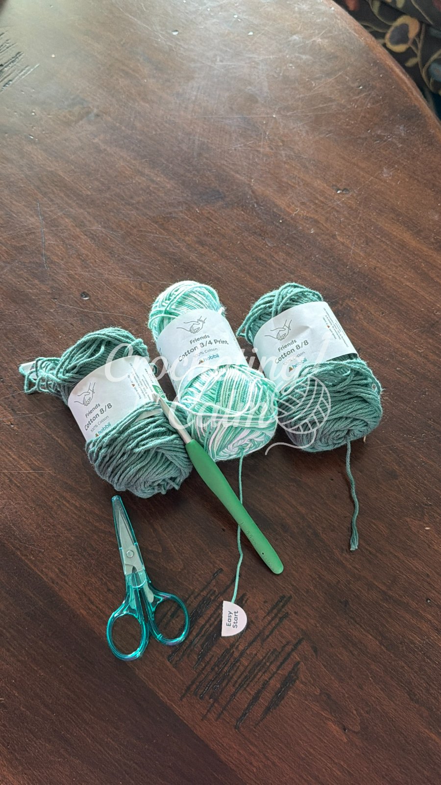 Green and white printed cotton yarn skeins with a green crochet hook and scissors laid out for a market bag project on a dark wooden table