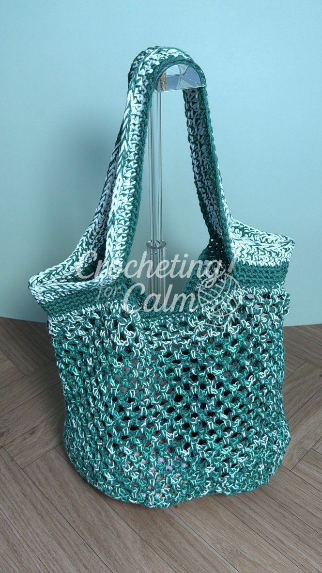 Sage green and white variegated mesh crochet market tote bag displayed hanging on an acrylic stand against a soft blue wall.