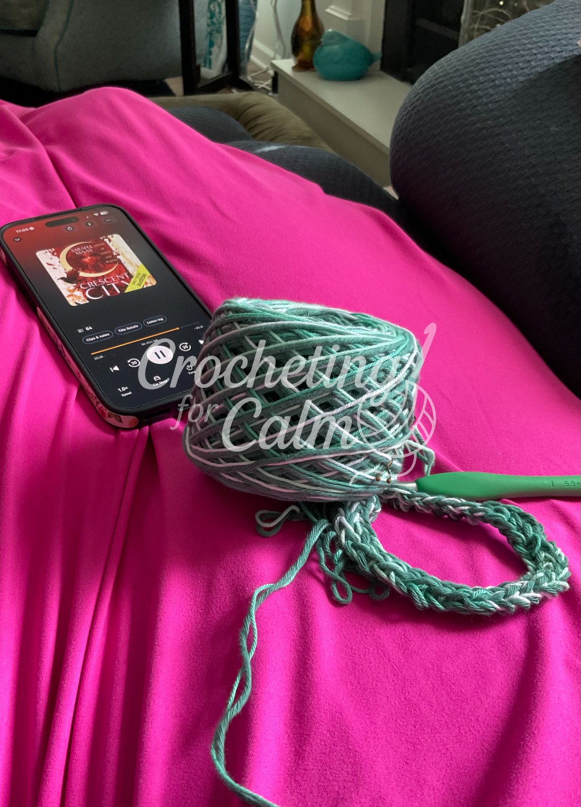 Sage green and variegated yarn cake resting on the lap of person wearing pink dress Also shows beginning crochet rounds and a green hook and an audio book open.