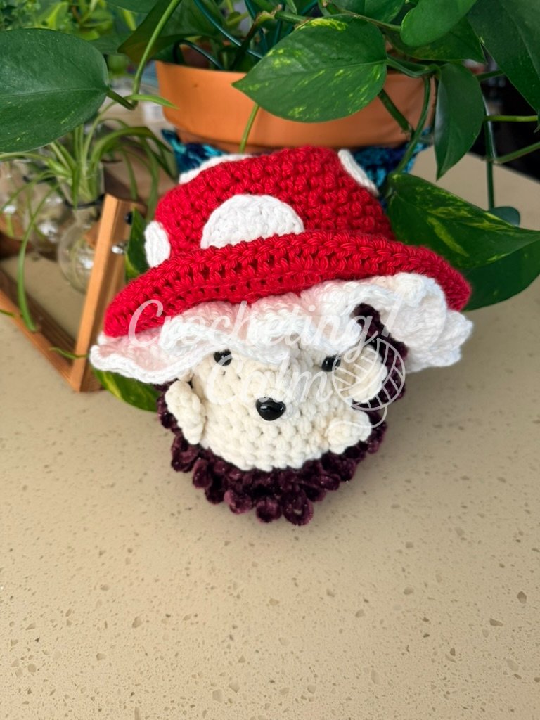 Red and white mushroom cap crochet baby hat featuring white spots and a ruffled cream brim, modeled on an amigurumi hedgehog.