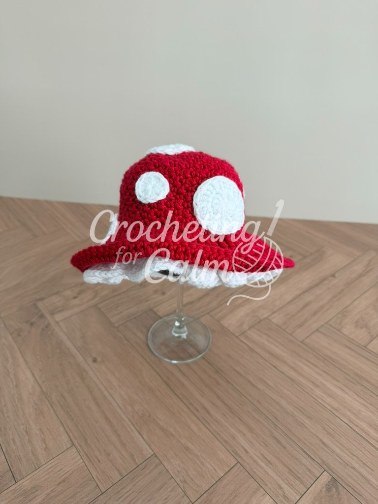 Red and white mushroom crochet baby hat with white polka dots and a ruffled underside, displayed upright on a clear wine glass over a wood surface.