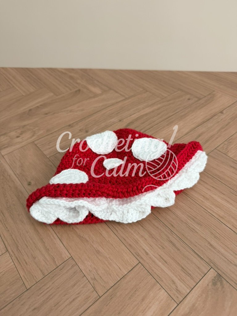 ed and white spotted mushroom cap crochet baby hat with a ruffled cream brim, laying flat on a light wood floor.