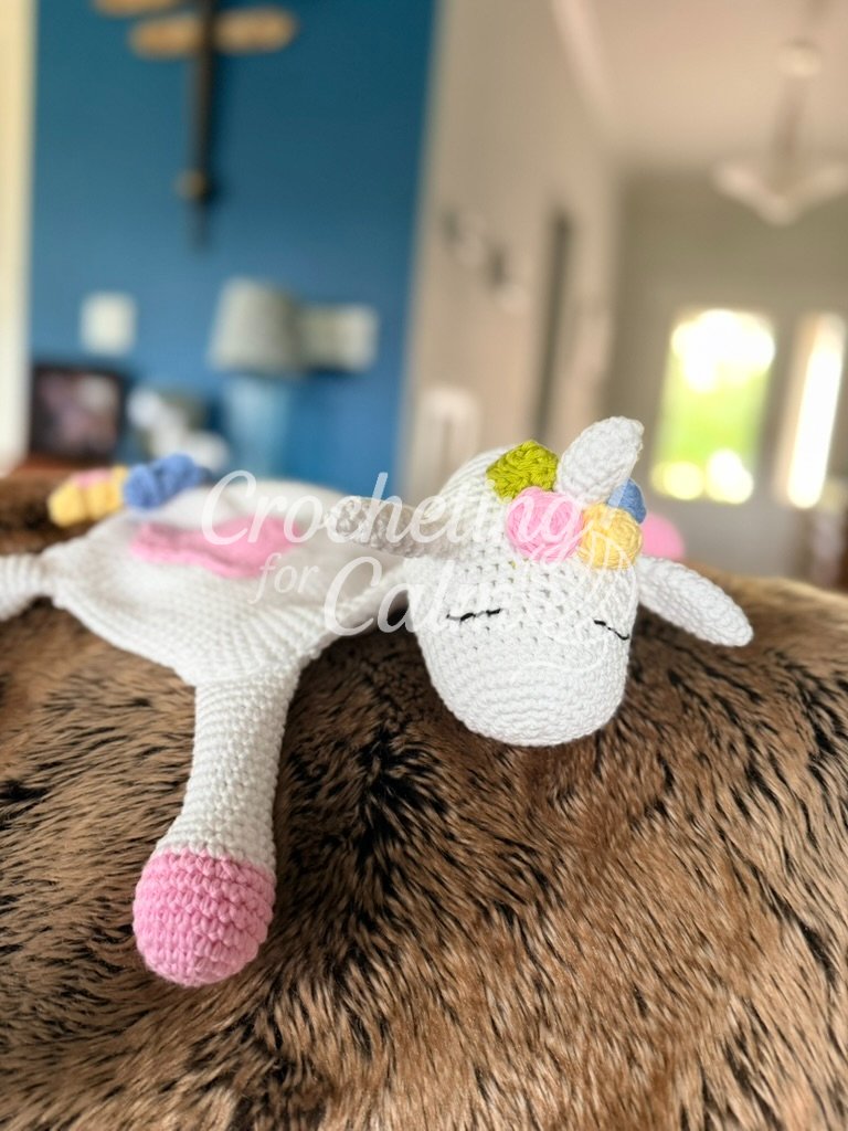Close-up profile view of a white unicorn crochet lovey showing a white horn, pastel green, pink, and yellow flower details, and embroidered eyelashes on a faux-fur surface.