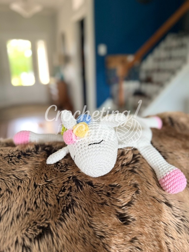 Front-facing view of a sleeping white unicorn crochet lovey with embroidered closed eyes and pastel floral mane accents, displayed on a brown textured blanket.