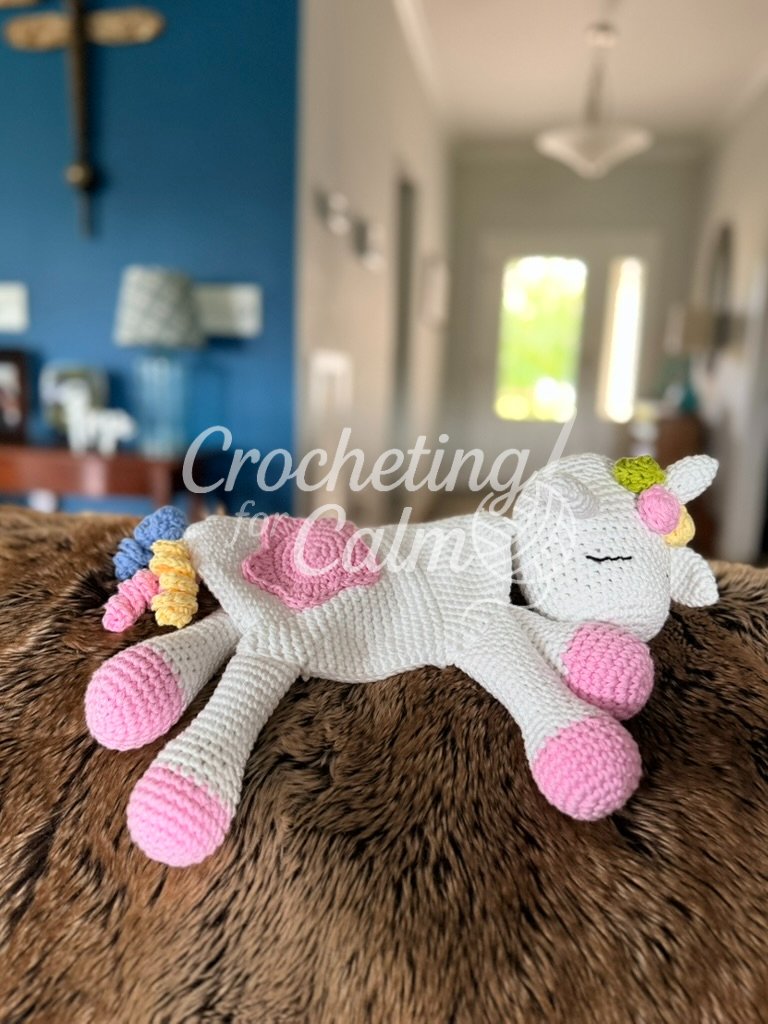 Full view of a white unicorn crochet lovey featuring a star-accented blanket body, colorful curled tail, and soft pink hooves, resting on a brown textured surface.