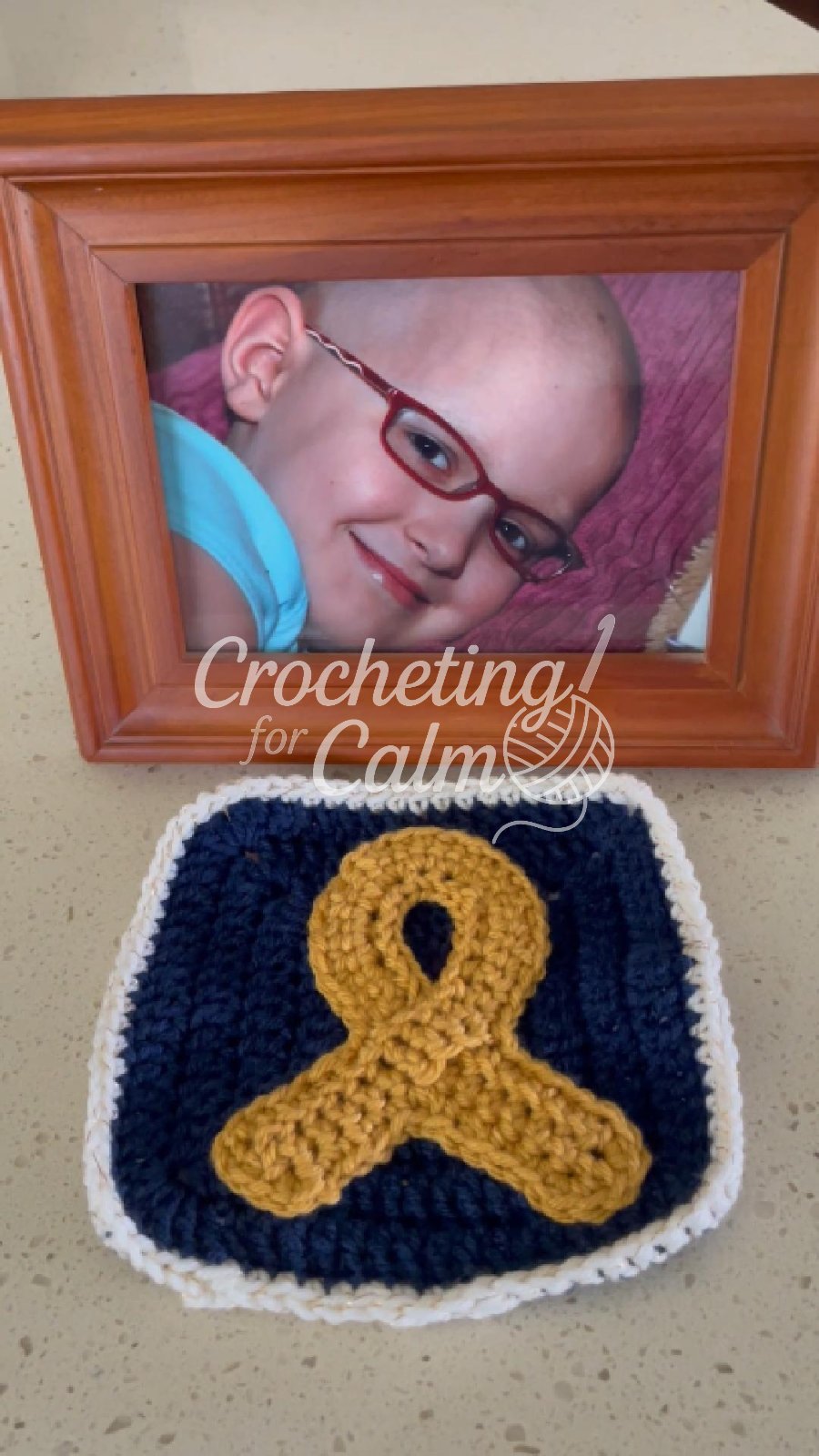 Handmade crochet granny square with a gold awareness ribbon for childhood cancer, stitched on navy blue with a white border by @crocheting4calm in front of a picture of her daughter, Colleen Moore who died of Osteosarcoma in 2008