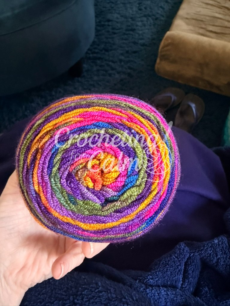 A top-down view of a vibrant, circular cake of 3-weight variegated yarn held in a hand, showing the rich color transitions from yellow to pink, purple, and green.