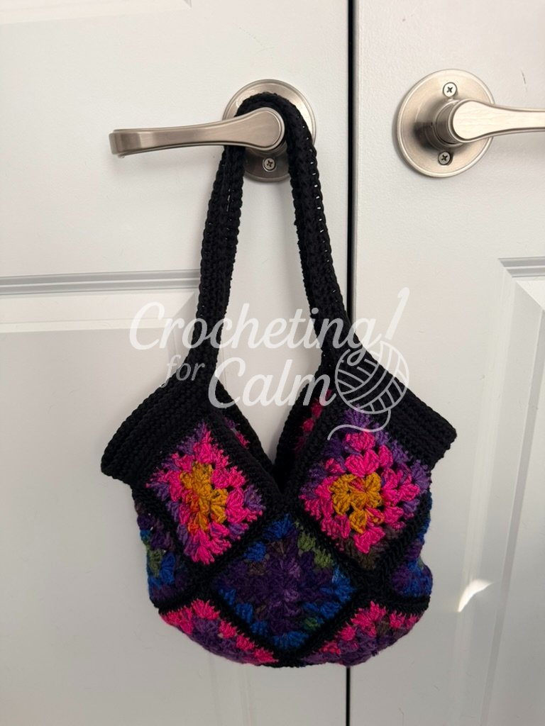 A finished black and colorful granny square 13 square tote bag hanging from a door handle bag has a wider top set of rows and handles because it was made with yarn that was too thick.