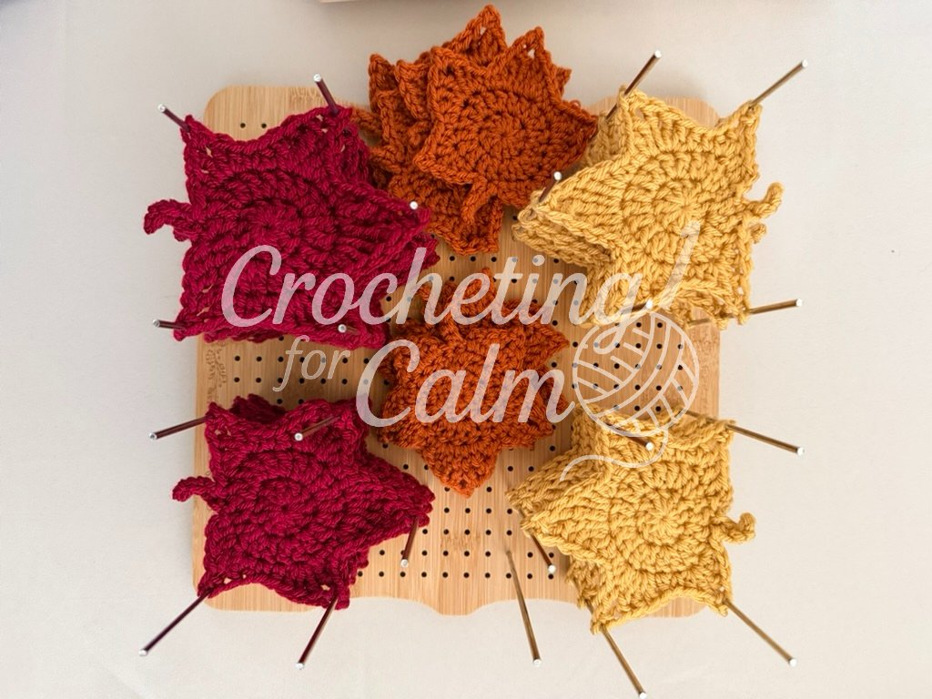 Close-up top-down view of a bamboo blocking board filled with handmade crochet autumn leaf appliques pinned in place, showing piles of burgundy, rust-orange, and mustard-yellow leave