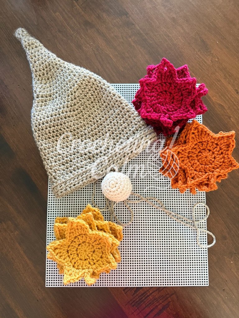 A handmade crochet gnome hat in grey yarn laid flat next to stacks of colorful autumn leaf appliques and a small white crocheted nose on a plastic grid sheet.