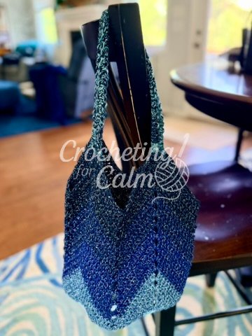 Dark blue and light blue variegated gradient crochet tote bag with a chevron stitch pattern and matching braided handles, hanging over the back of a dark wooden chair on a patterned blue rug.