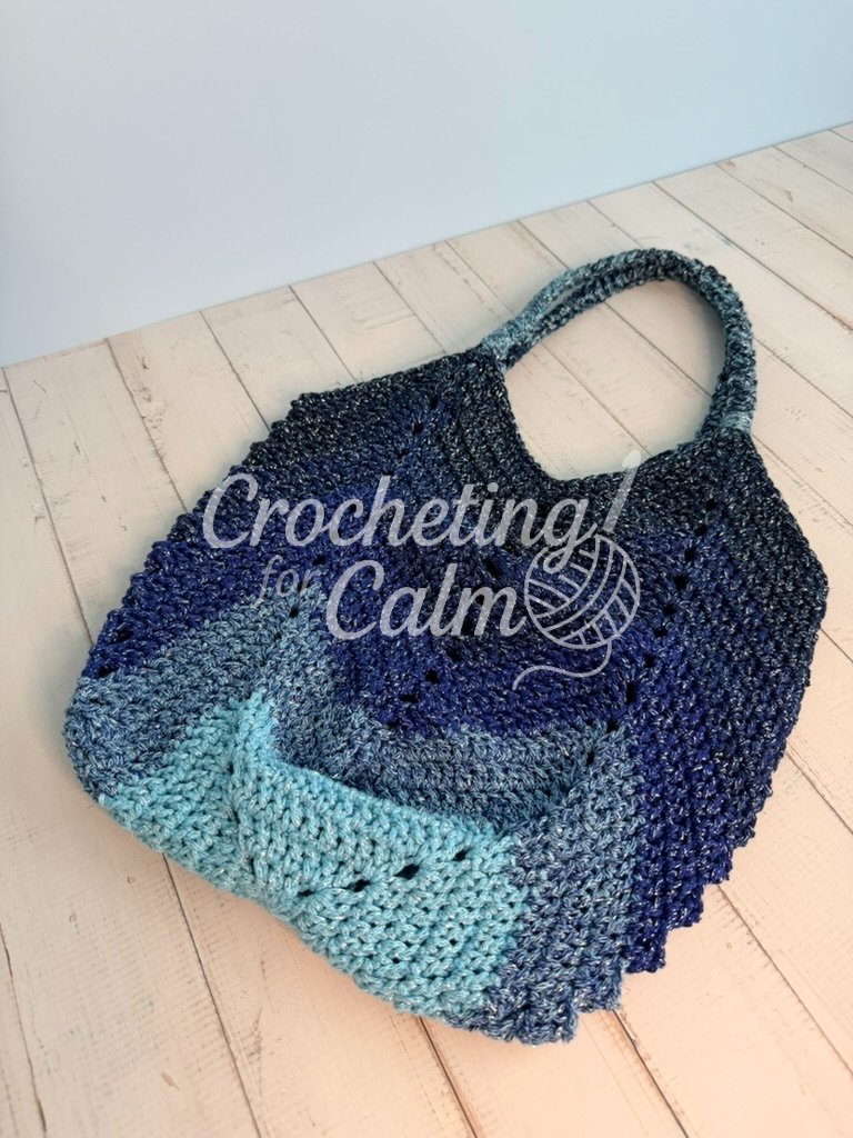 Dark blue and light blue variegated gradient crochet tote bag with matching textured braided shoulder straps, angled on a white wooden surface