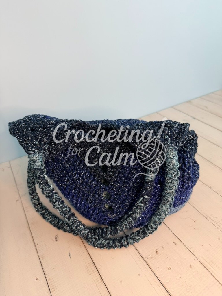 Dark navy blue and light blue variegated gradient crochet tote bag featuring a textured chevron stitch pattern and braided shoulder handles, resting on a white wooden surface.