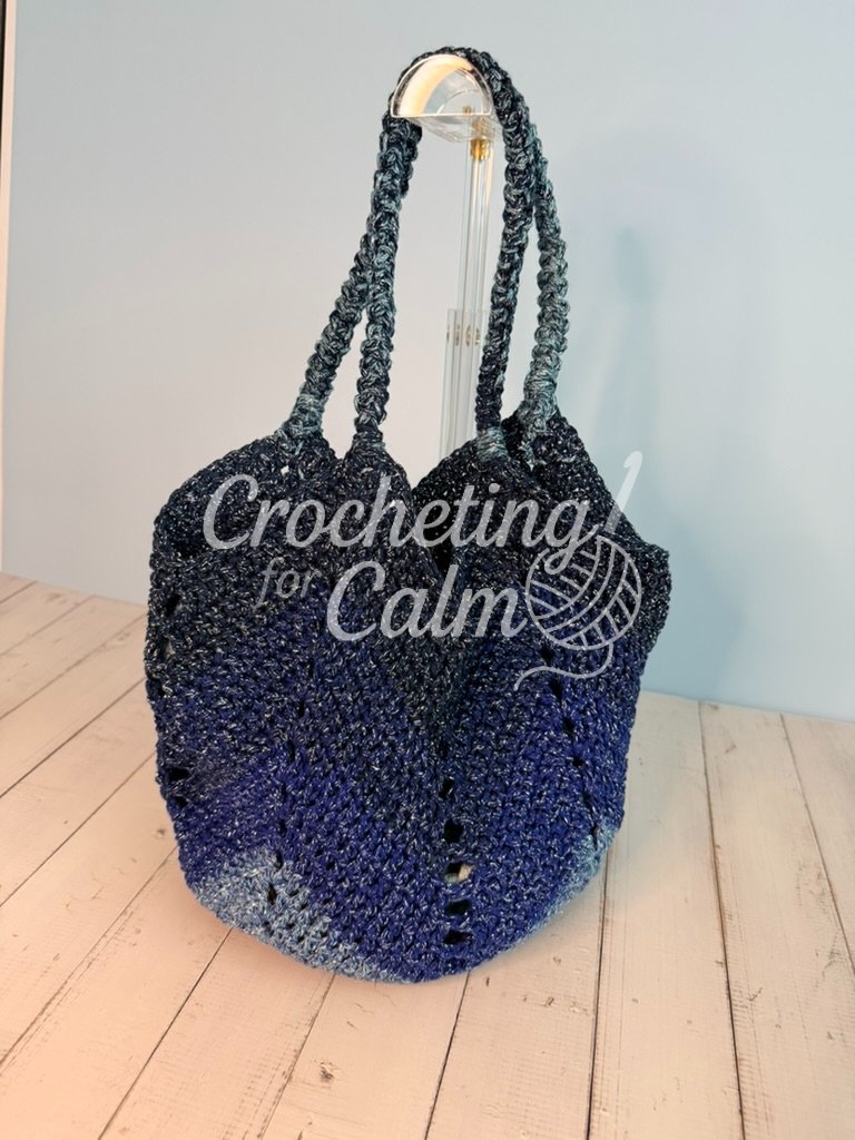 Dark blue, royal blue, and light blue variegated gradient crochet tote bag featuring a chevron stitch pattern and braided handles, hanging from a clear acrylic stand against a light blue wall.