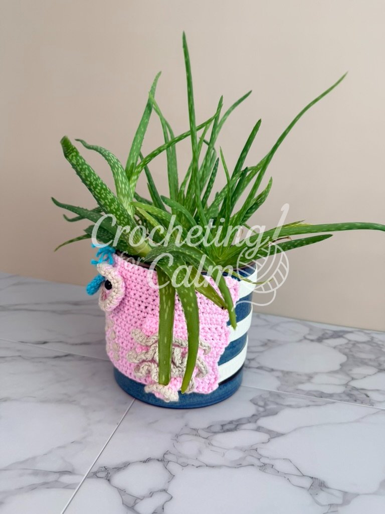 Side profile view showing how the pink crochet owl cozy wraps around the blue planter, highlighting the cream textured wing and the three wide white elastic bands stretched across the back.