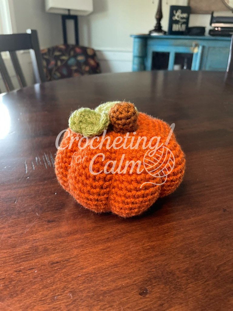 Front-facing view of an orange crochet pumpkin with a brown stem and light green leaves, resting on a dark wood table with a chair and blue cabinet in the background.