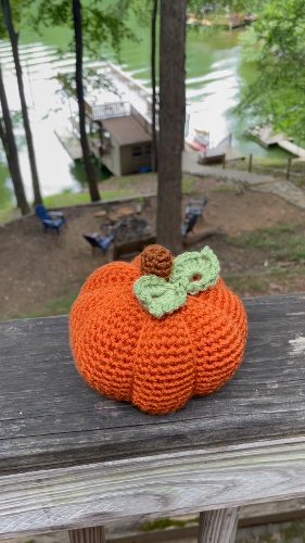 Orange crochet pumpkin with a brown textured stem and a light green leaf, resting on a weathered wooden deck with a lake, dock, and trees in the background.