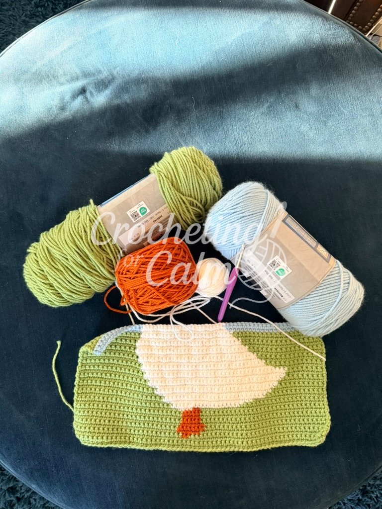 Flat lay of crochet supplies on a dark textured round surface, featuring skeins of sage green and soft blue yarn, a small orange yarn cake, a ball of white yarn, a purple crochet hook (size 4mm), and a partially worked tapestry crochet panel depicting a white duck on a green field.