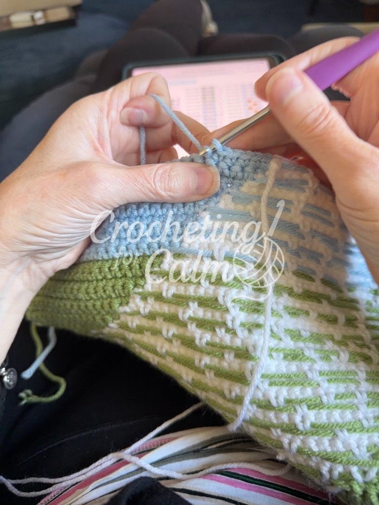 lose-up of hands working on a tapestry crochet project, with a digital chart visible on a tablet in the background and a purple-handled 4mm crochet hook inserting into stitches atop a blue sky and textured green landscape.