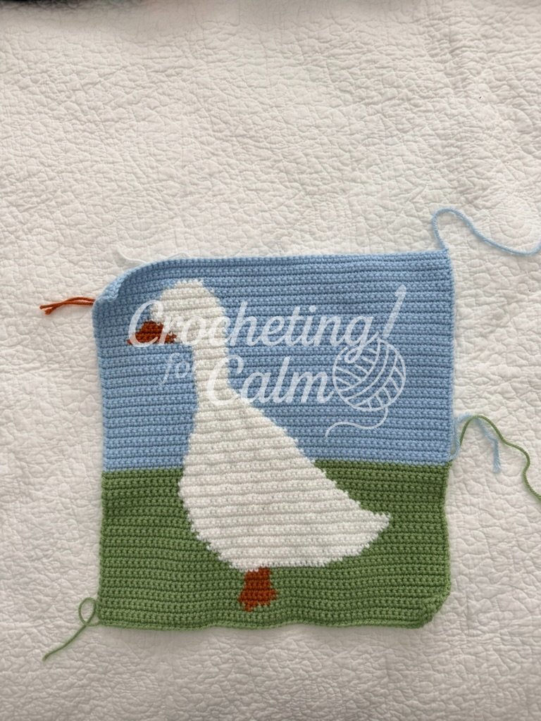 A completed tapestry crochet panel laid out flat on a textured white surface, featuring a white duck with an orange bill and feet standing on a green grass section against a light blue sky background.