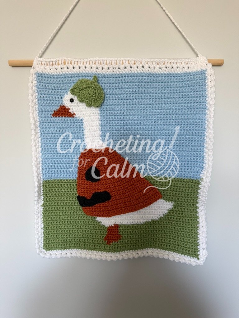 White, orange, and green crochet tapestry wall hanging featuring a detailed goose wearing a green beret and an orange sweater with black accents, displayed hanging on a wooden dowel against a neutral wall.