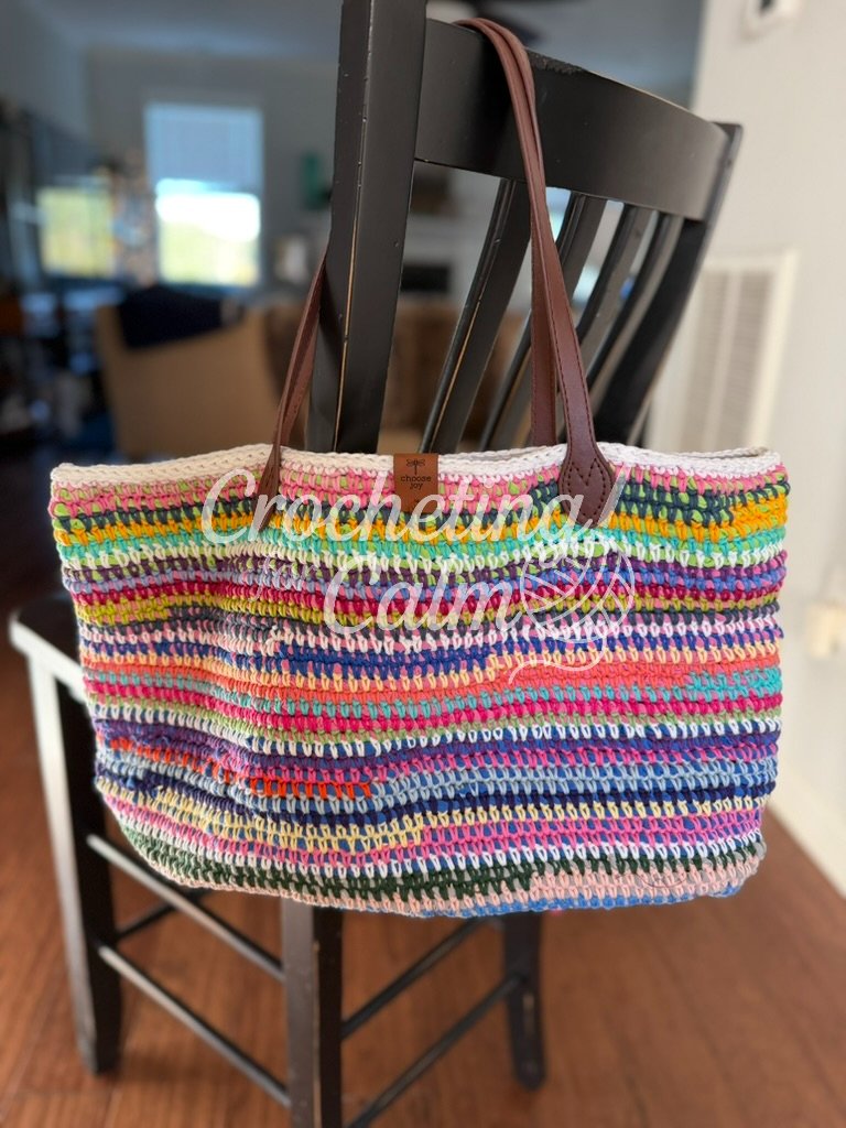 Multi-colored textured crochet tote bag (made with t-shirt yarn wrapped with scrap cotton yarn) with dark brown leather handles, hanging from a black wooden chair against a light-filled background.